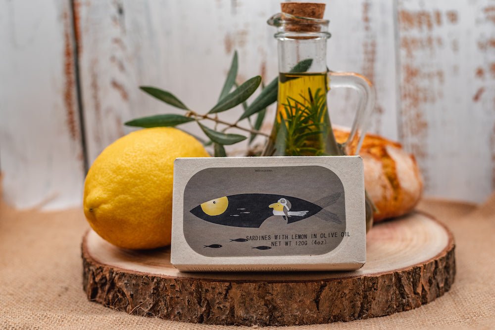 Ein Stück Seife mit Illustrationsverpackung für Olivenöl wird auf einer Holzscheibe präsentiert, begleitet von einer frischen Zitrone, einem Olivenzweig und einer Dose Sardinen in Olivenöl mit Zitrone von Jose Gourmet vor einem rustikalen Holzhintergrund.
