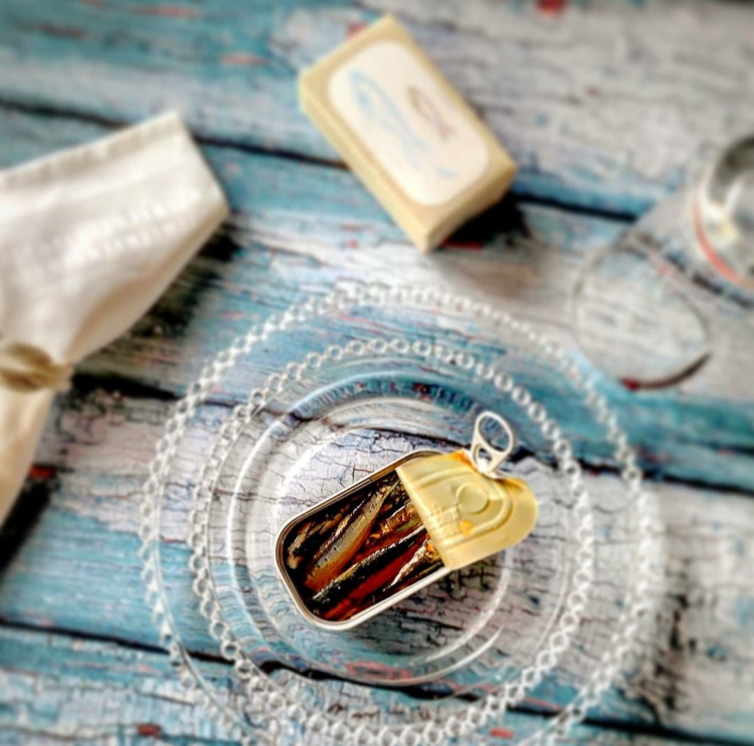 Eine Dose mit kleinen, scharfen Sardinen in Olivenöl von Jose Gourmet auf einer Spitzenunterlage, dazu Wasser und ein schlichtes Tischgedeck.