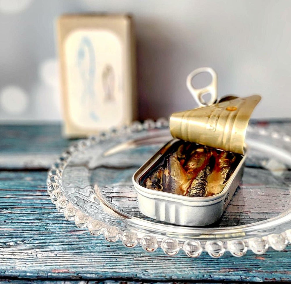 Eine geöffnete Blechdose mit kleinen, scharfen Sardinen in Olivenöl von Jose Gourmet auf einem dekorativen Glasteller mit unscharfem Hintergrund auf einer Holzoberfläche.