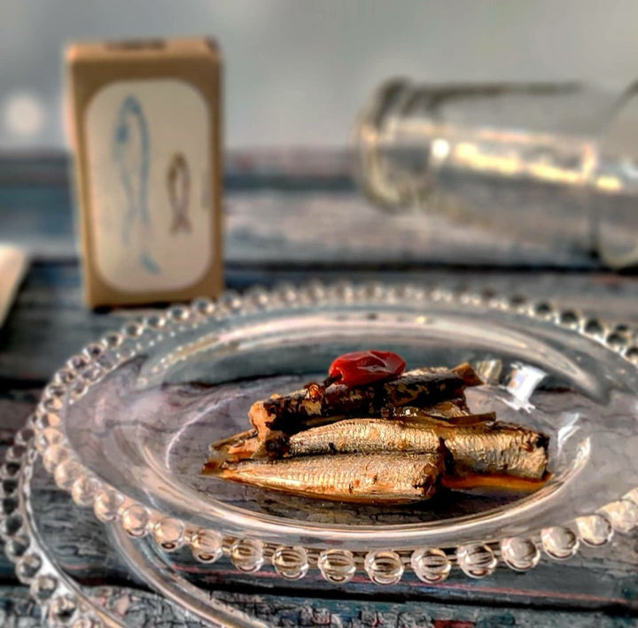 Auf einem Glasteller stehen mehrere kleine Jose Gourmet Pikante Sardinen in Olivenöl mit Garnitur auf einem Holztisch, im Hintergrund ein Salzstreuer und eine umgekippte Glasflasche.