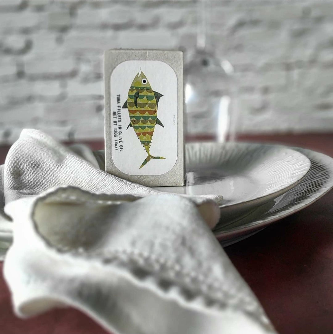 Eine Spielkarte mit einer Fischillustration, symbolisch für Jose Gourmet Thunfischfilets in Olivenöl, wird an eine durchsichtige Flasche gelehnt, vor einem Teller mit einem gefalteten Stofflappen.