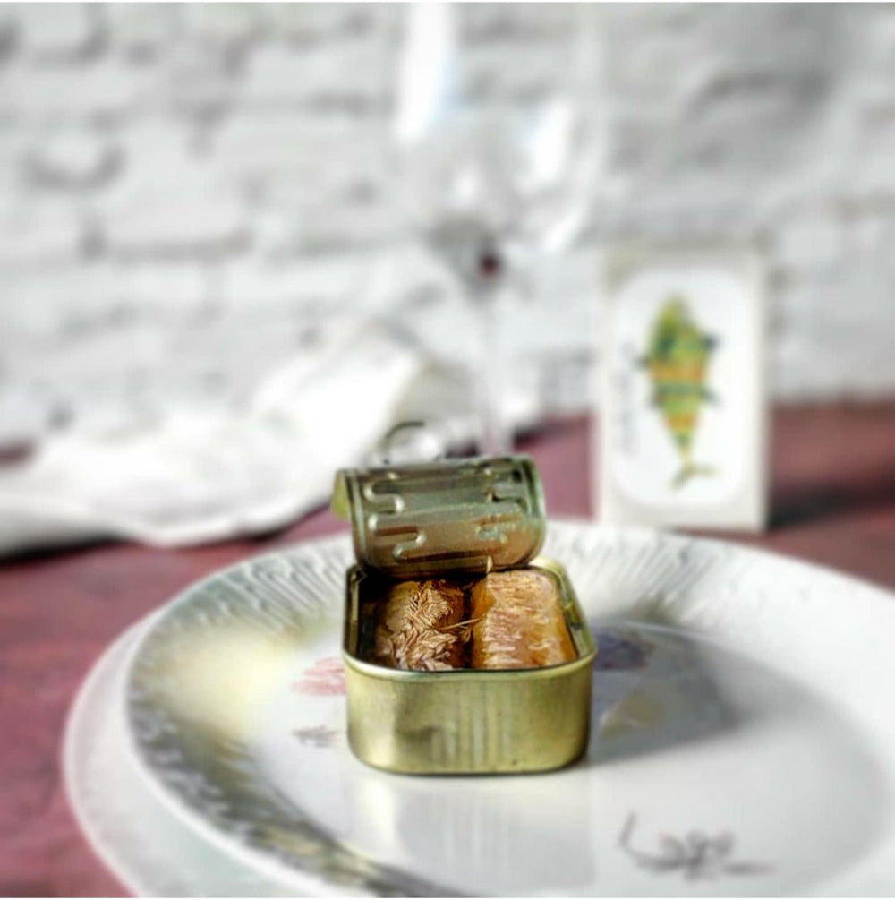 Eine teilweise geöffnete Blechdose Jose Gourmet Thunfischfilets in Olivenöl, mit ein paar sichtbaren Sardinen, auf einem dekorativen Teller mit verschwommenem Hintergrund.