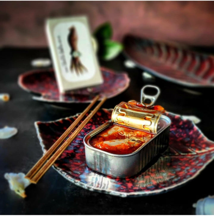 Eine offene Dose gefüllte Kalmartuben (Tintenfisch) in Ragoutsauce von Jose Gourmet auf einem dekorativen Teller mit Stäbchen zur Seite, vor einem verschwommenen Hintergrund, der Anschein