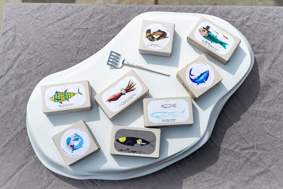 Eine Sammlung von illustrierten Untersetzern mit Designs des maritimen Lebens, angeordnet auf einem weißen geschwungenen Tisch neben einem Metallspachtel und gefüllten Kalmartuben (Tintenfisch) in Ragoutsauce von Jose Gourmet.