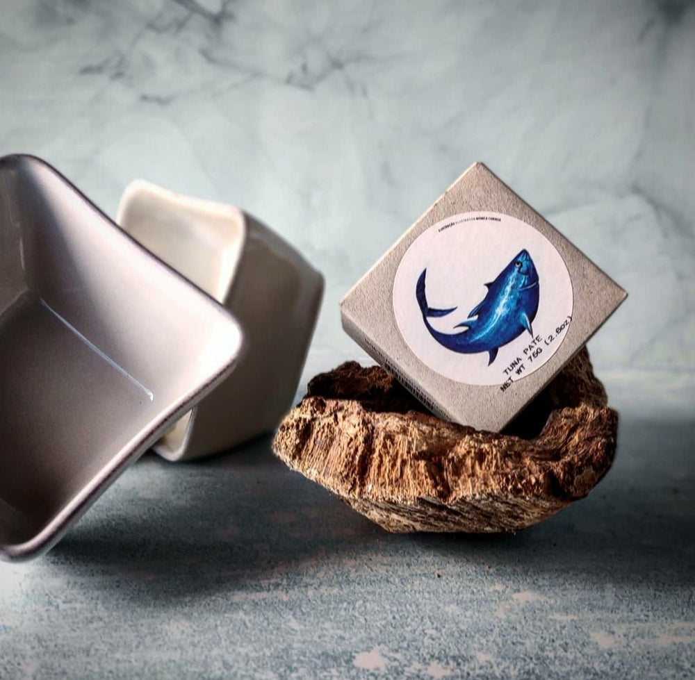 Eine Thunfisch-Mousse mit einer Illustration und einem Text aus blauen Federn wird auf einem naturbraunen Objekt mit rauer Textur balanciert und erinnert an die Essenz von Jose Gourmet, mit einem umgekippten hellbeigen Tablett.