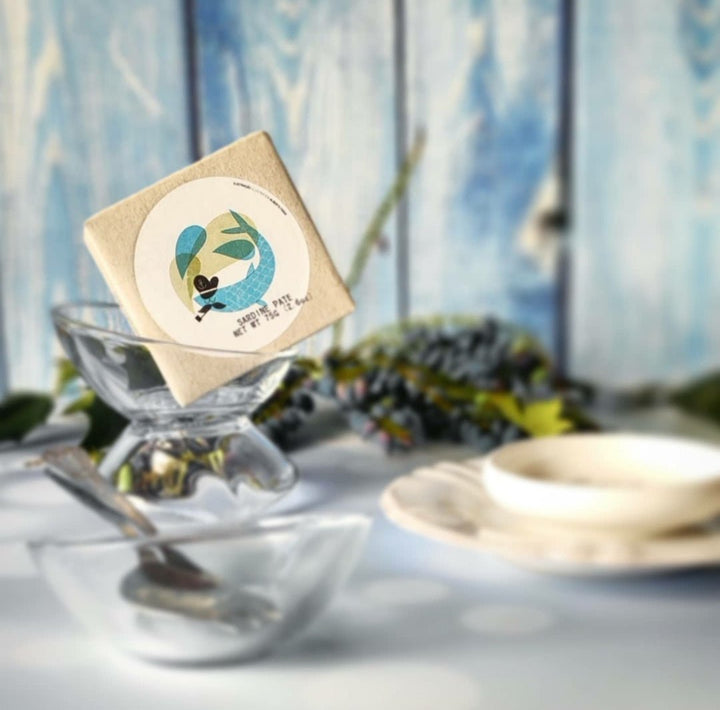 Auf einer durchsichtigen Glasschale, Teil eines Tischgedecks mit rustikalem blauem Hintergrund, liegt ein kleines quadratisches Päckchen mit einer Vogelillustration, das Jose Gourmets portugiesischen Atlantikküstenfang Sardinenmousse enthält.