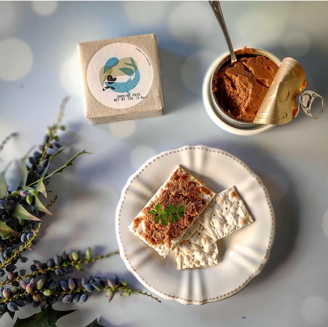 Eine geöffnete Dose Jose Gourmet Sardinenmousse neben Crackern mit Pastete und Kräutern, dazu ein dekorativer Zweig und eine Papierserviette mit einer Pinguinillustration.