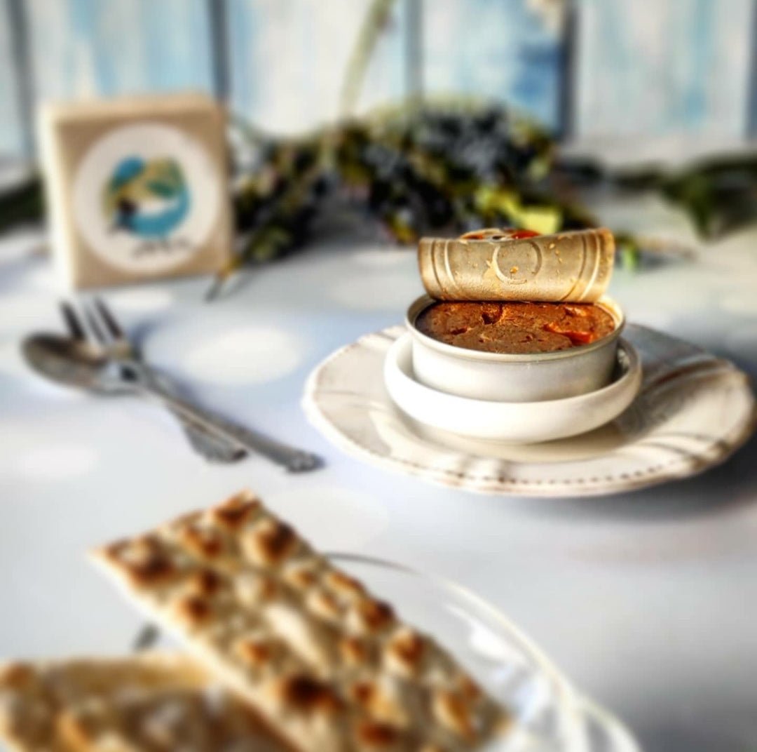 Ein fein präsentiertes Gericht mit Sardinenmousse von Jose Gourmet auf einem weißen Teller mit knuspriger, goldener Kruste, platziert auf einem Restauranttisch mit verschwommenem Hintergrund mit Besteck und dekorativen Elementen.