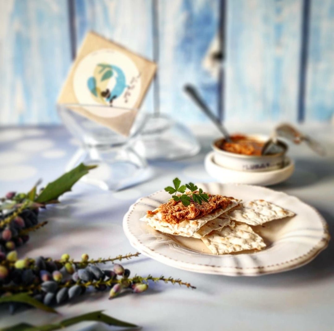 Ein Teller mit Crackern und einem Sardinen-Mousse-Aufstrich von Jose Gourmet, garniert mit einem Zweig Petersilie, einer Schüssel mit zusätzlichem Aufstrich, einem Messer und einer dekorativen Pflanze vor einem blauen Holzhintergrund.