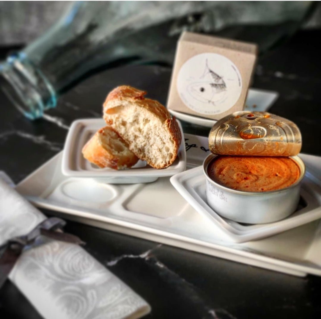 Eine einfache, aber elegante Bordmahlzeit mit einem geteilten Brötchen, einem Behälter mit Piri-Piri-Aufstrich, einer kleinen Dose Kaviar und dem dazugehörigen Besteck, alles arrangiert auf Makrelen-Mousse von Jose Gourmet.