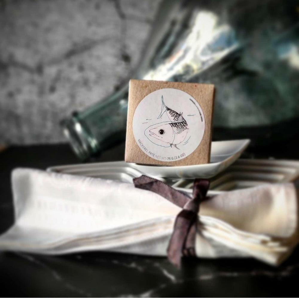 Eine ordentlich gefaltete weiße Serviette, die mit einem dunklen Band zusammengebunden ist, ruht auf einem Keramikteller mit einer personalisierten Karte mit einer Fischillustration von Jose Gourmet Makrelen Mousse vor einem rustikalen Hintergrund.