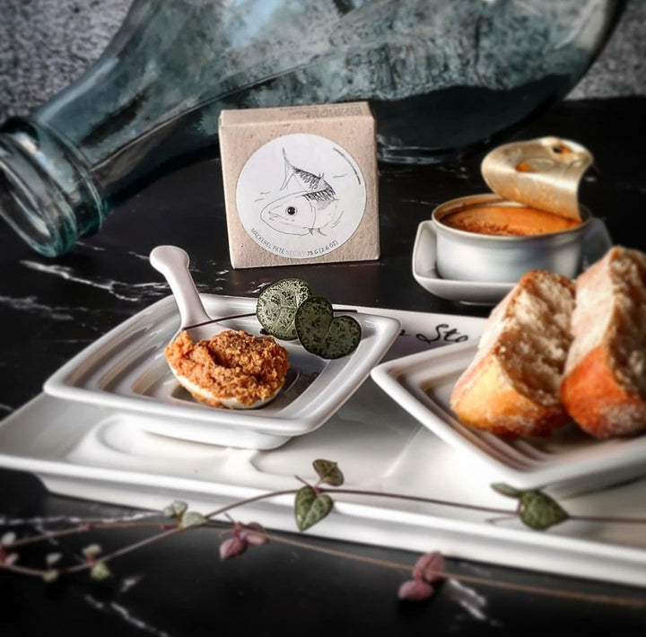 Eine Stilllebenkomposition einer Frühstücksszene mit einer Tasse Kaffee mit Schaumkunst, einer kleinen Schüssel mit Makrelen-Mousse von Jose Gourmet, Brotscheiben und dekorativen grünen Blättern auf einer dunklen Oberfläche daneben.