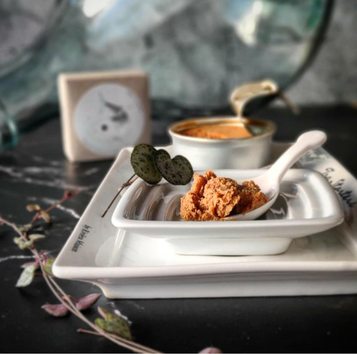 Ein Löffel cremiges Jose Gourmet Makrelen Mousse mit knusprigem Belag ruht auf einem eleganten weißen Teller, begleitet von einer vollen Desserttasse und einer Kaffeetasse im Hintergrund.