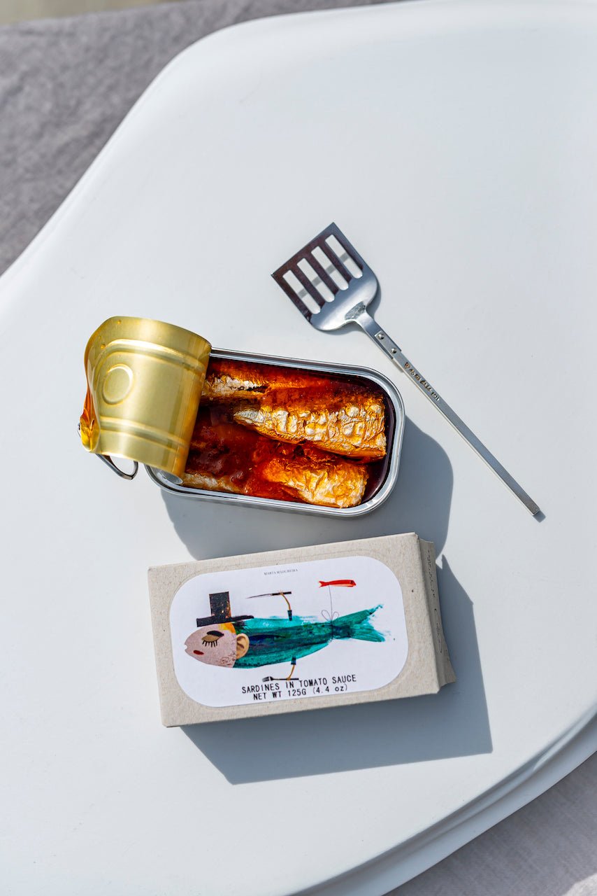 Eine Dose Fischkonserven in Tomatensauce geöffnet und auf einer weißen Oberfläche platziert, begleitet von einem Sardinenheber aus 925er Sterlingsilber von Maritime Zeitkapseln.