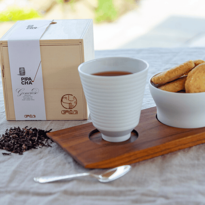 Eine Tasse Portwein Tee – Pipa Cha wird auf einer hölzernen Untertasse mit daneben verstreuten losen Teeblättern, begleitet von einer Schachtel Tee und einem Teller Kekse, auf einem Tisch im Freien mit einem Platz gestellt.