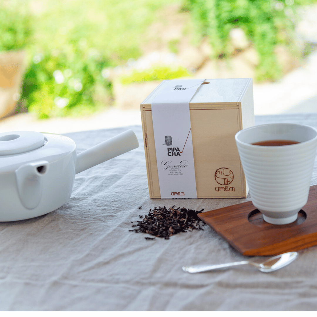 Eine Schachtel Portwein-Tee – Pipa Cha neben losen Teeblättern auf einem Holzbrett, mit einer weißen Teekanne und einer gefüllten Tasse auf einem mit Leinen bedeckten Tisch.