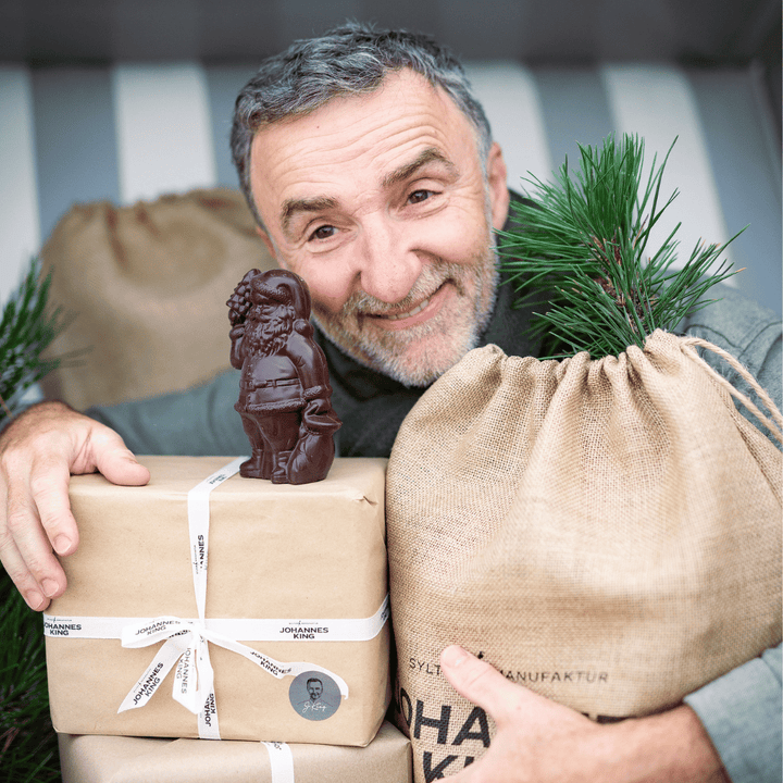 Ein Mann lächelt, während er einen Geschenkbeutel im Weihnachtsdesign der Sylter Manufaktur Johannes King, eine umweltfreundliche Weihnachtsverpackung und einen Leinensack neben einem Tannenzweig hält.