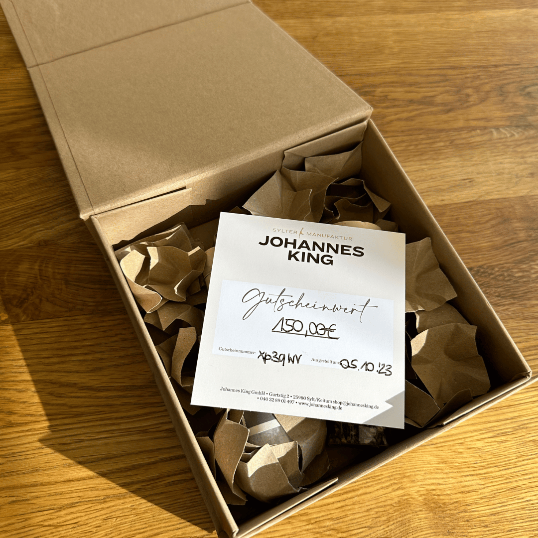 Ein Geschenkbox-Gutschein der Sylter Manufaktur Johannes King liegt auf zerknittertem Packpapier in einem offenen Karton und wird von natürlichem Licht beleuchtet. Das Zertifikat ist handschriftlich personalisiert und weist einen Wert von 150 Euro aus.