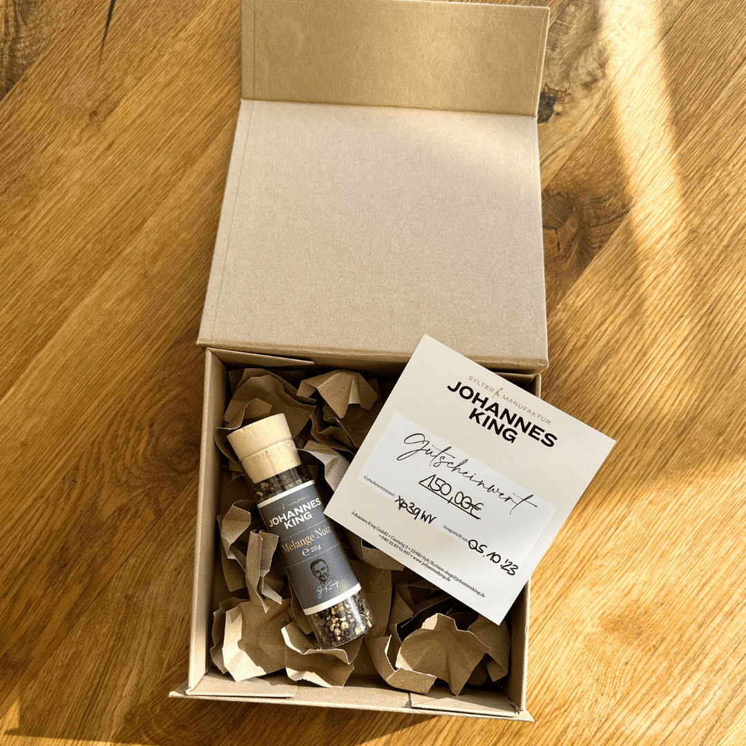 Eine offene Geschenkbox mit Melange Noir auf einer Holzoberfläche, darin eine Flasche mit Etikett und eine Visitenkarte mit Handschrift darauf, eingebettet in zerknittertes Packpapier. Markenname: Sylter Manufaktur Johannes King