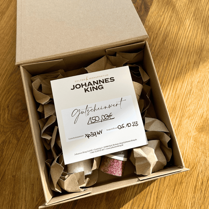 Eine geöffnete Geschenkbox mit Salz-Duo mit Knitterpapierfüllung enthält einen Geschenkgutschein der Sylter Manufaktur Johannes King.