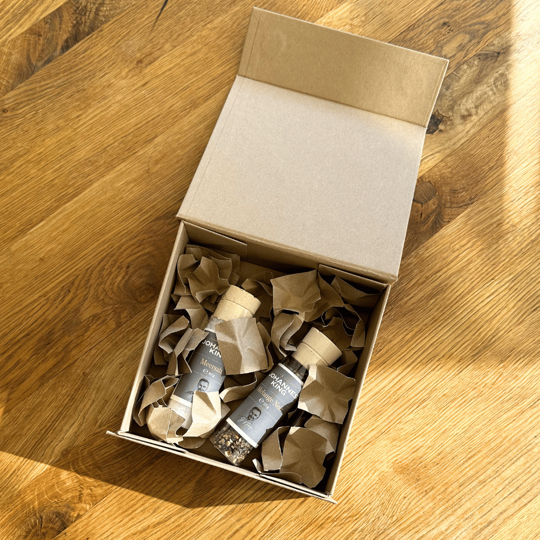 Eine Geschenkbox mit Sylter Meersalz und Pfeffer der Sylter Manufaktur Johannes King liegt auf einem Holzboden im Sonnenlicht.