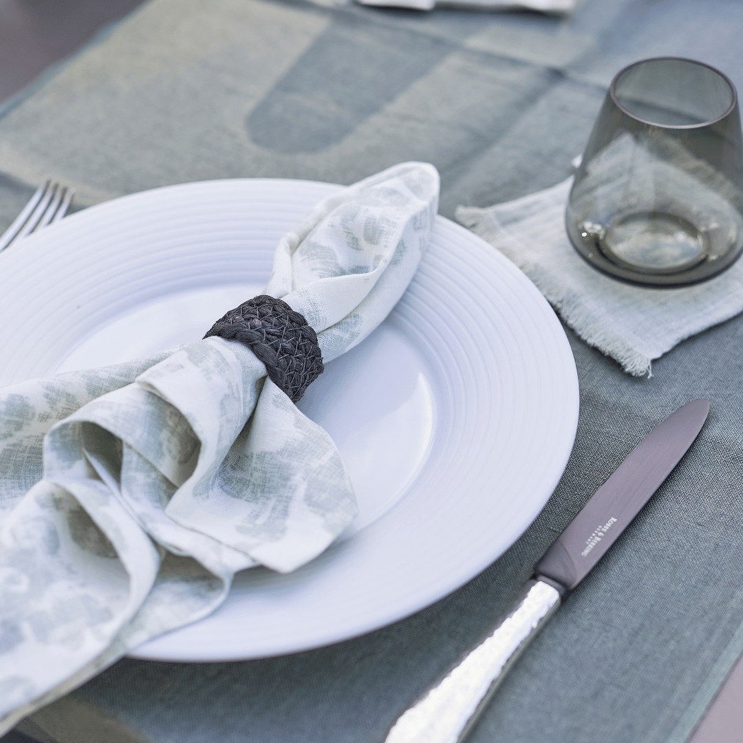 Ein ordentlich gedeckter Tisch mit einem weißen Teller, einer Serviette Dorothea Waydsch von Dorothea Waydsch, einem dunklen Serviettenring, Besteck und einem leeren Glas auf einer blauen Tischdecke