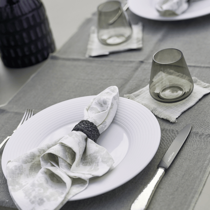 Ein ordentlich arrangierter Esstisch mit weißen Tellern, Besteck, gemusterten Servietten und klaren Gläsern, gedeckt auf einer grauen Tischdecke von Tischwäsche-Set Dorothea Waydsch.