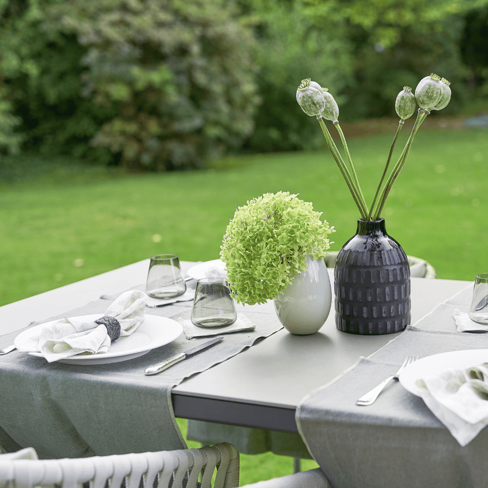 Ein Tischgedeck im Freien mit einer weißen Vase mit einem grünen Blumenarrangement, einer dunkelfarbigen strukturierten Vase mit Mohnkapseln, klaren Gläsern und gefalteten Servietten auf einem grauen Tischwäsche-Set Dorothea Waydsch von Dorothea Waydsch.