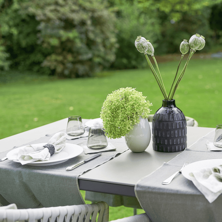 Ein Tischgedeck im Freien mit einer weißen Vase mit einem grünen Blumenarrangement, einer dunkelfarbigen strukturierten Vase mit Mohnkapseln, klaren Gläsern und gefalteten Servietten auf einem grauen Tischwäsche-Set Dorothea Waydsch von Dorothea Waydsch.