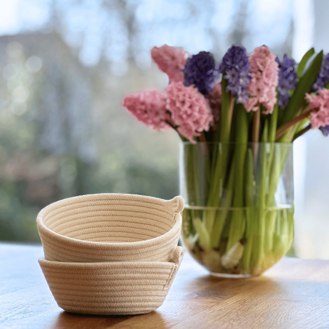 Ein Stapel beiger Aufbewahrungskörbe vom Typ „Mia Mélange Baumwollkorb, klein“ (T 15 cm x H 5 cm) steht auf einem Tisch im Vordergrund, im Hintergrund steht eine Glasvase mit blühenden rosa und lila Hyazinthen.