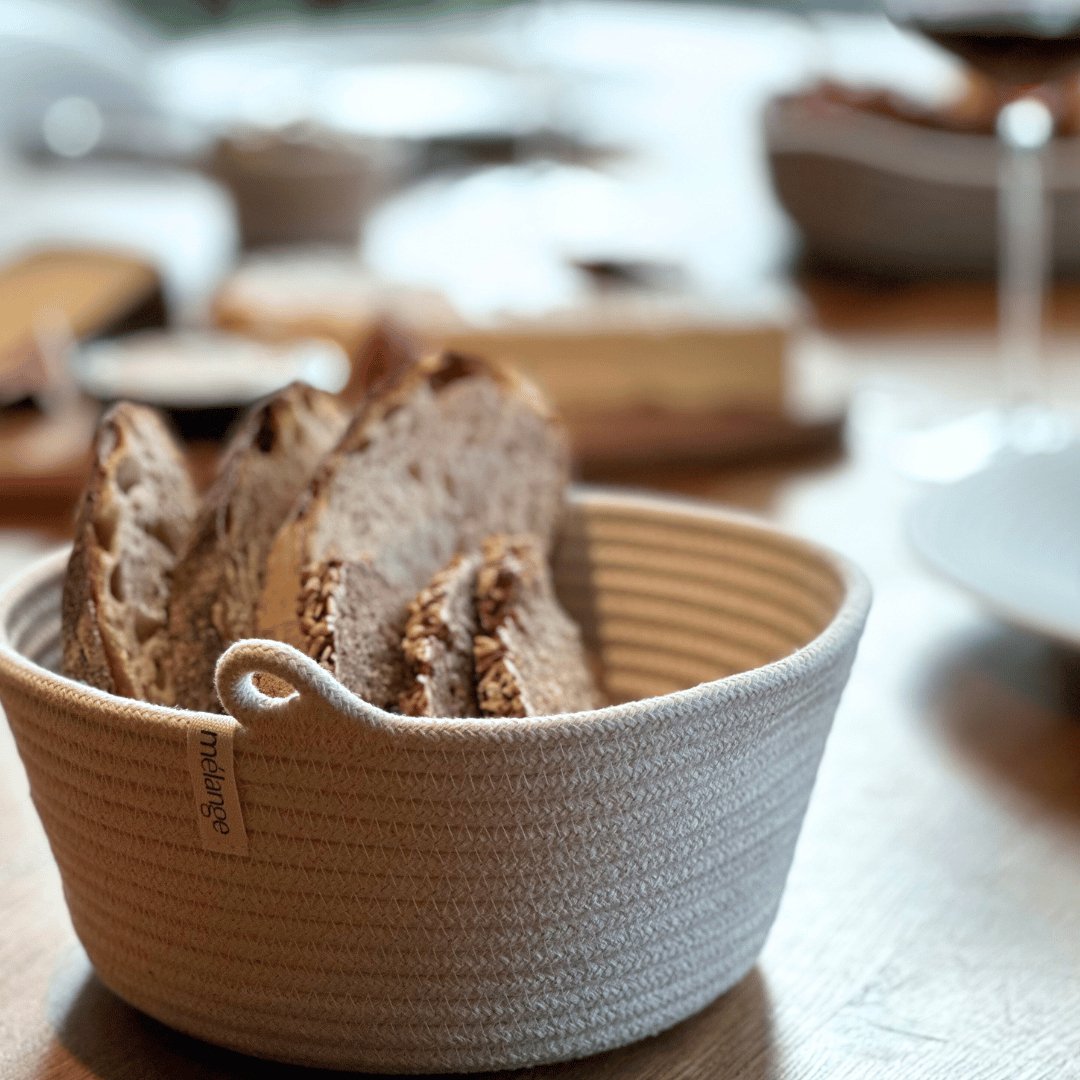 Ein Korb mit geschnittenem Brot auf einem Holztisch mit verschwommenem Hintergrund, der auf zusätzliches Geschirr schließen lässt, darunter Mia Mélanges Baumwollkorb, klein (D 15 cm x H 5 cm).