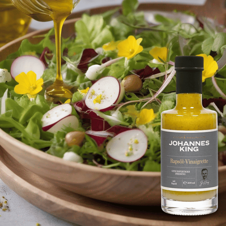 Ein frischer Salat mit essbaren Blüten und eine Flasche Vinaigrette-Duo der Marke Sylter Manufaktur Johannes King, dazu wird Öl aus der Höhe eingegossen.