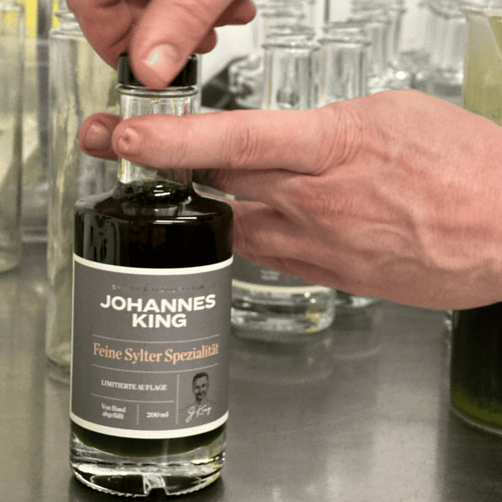 Eine Hand öffnet eine kleine Glasflasche mit der Aufschrift „Sylter Manufaktur Johannes König Schnittlauchöl“ auf einer Küchenarbeitsplatte, im Hintergrund verschiedene Glaswaren.