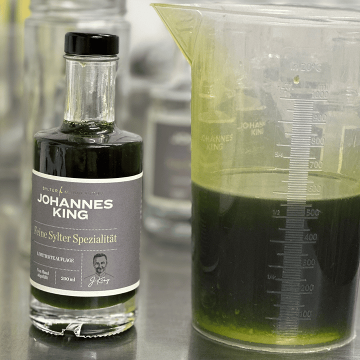 Eine Flasche des Gourmet-Lebensmittelprodukts Kings Schnittlauchöl der „Sylter Manufaktur Johannes King“ neben einem Messbecher mit einer grünen Flüssigkeit.