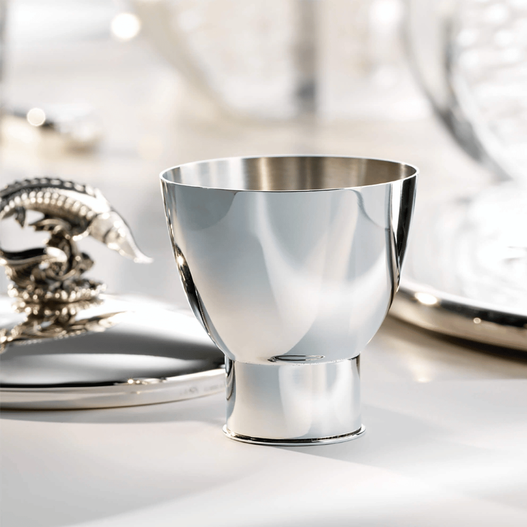 Robbe & Berking Schnapsbecher Sylt Edition auf einem Tisch mit spiegelnder Oberfläche und elegantem Geschirr im Hintergrund, das exquisite Silberschmiedekunst präsentiert.