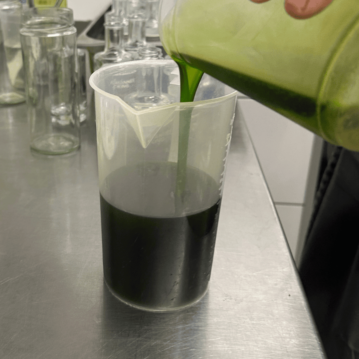 Eine Person gießt ein grünes Sylter Manufaktur Johannes King Schnittlauchöl mit Salz & Pfeffer aus einem Behälter in einen durchsichtigen Messbecher auf einer Edelstahloberfläche.